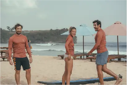 Three people wearing rash guards smile on a sandy beach with a surfboard and ocean waves in the background