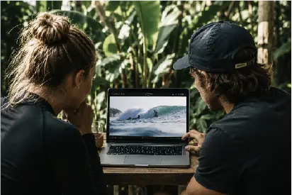 Two people outdoors looking at a laptop screen displaying a video of a surfer on a wave