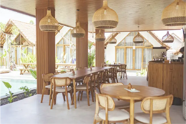 Semi-outdoor dining area with wooden tables and woven pendant lights overlooking a pool and resort bungalows