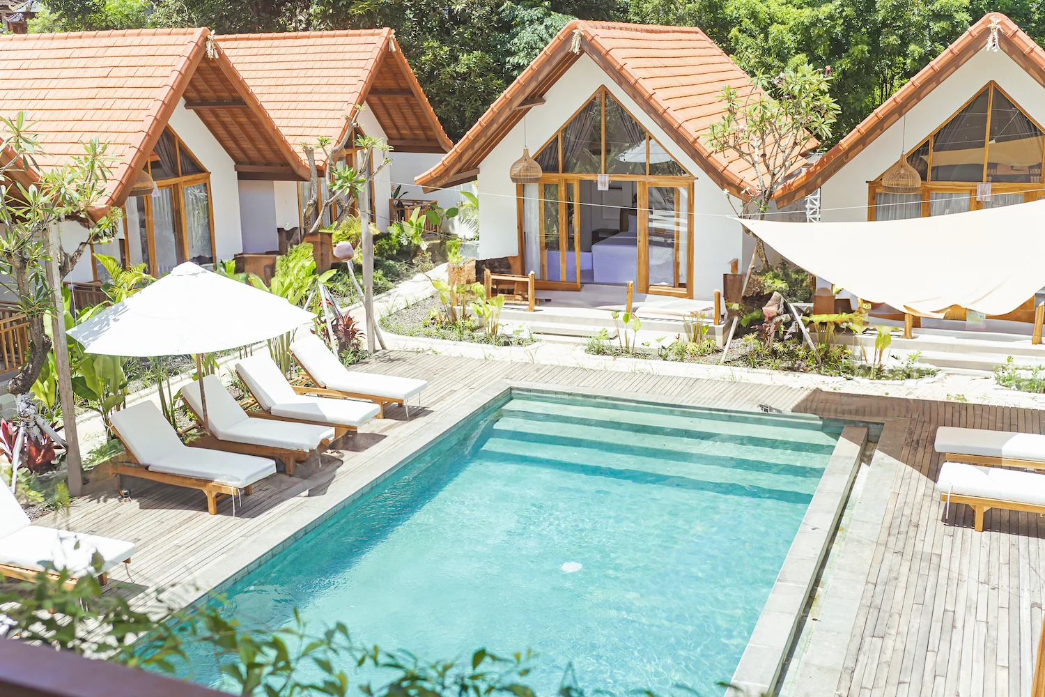 Swimming pool and wooden deck with lounge chairs in front of white A-frame cottages with orange-tiled roofs