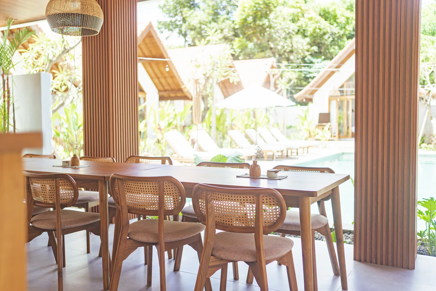 Wooden dining table and chairs in an open-air pavilion overlooking a resort pool and tropical villas