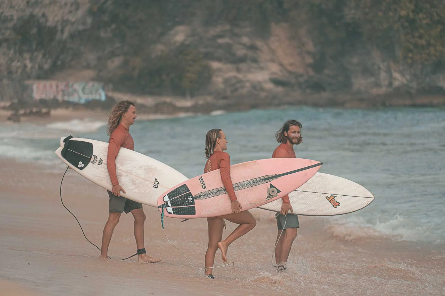 Three surfers carrying surfboards walk along a sandy beach shoreline